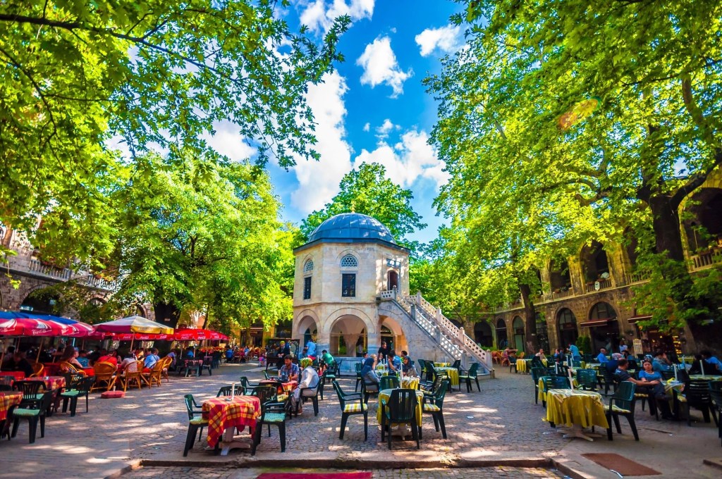 Yeşil ağaçlarla çevrili, açık hava kafeleri olan bir meydan. Ortada tarihi bir yapı ve kalabalık bir atmosfer.