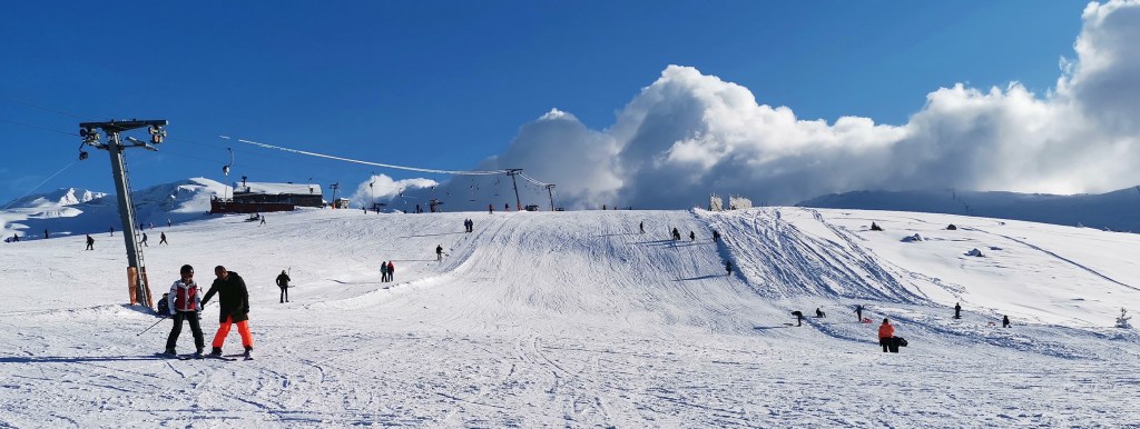 Karla kaplı bir dağın zirvesinde kayak yapan insanlar, arka planda teleferik ve karlı manzara.