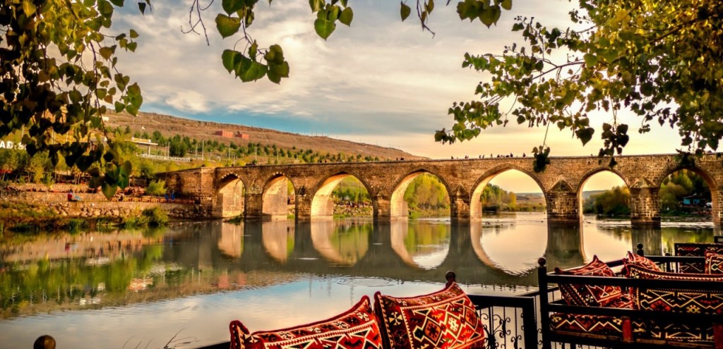 Bir nehir üzerinde tarihi bir taş köprü, suyun yansımasıyla birlikte görüntüleniyor. Köprünün etrafında doğal bir manzara ve oturma alanları bulunuyor.