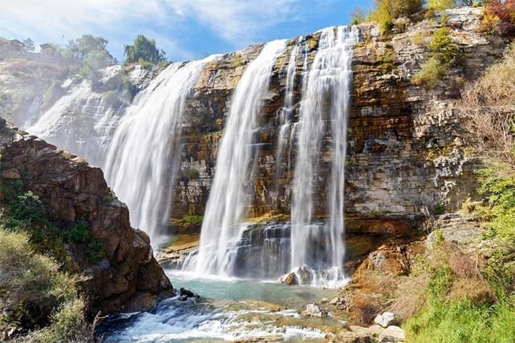 Şelale ve çevresindeki doğal manzara. Akışkan suyun kayalıklardan düştüğü ve etrafında yeşil bitkilerin bulunduğu bir görüntü.