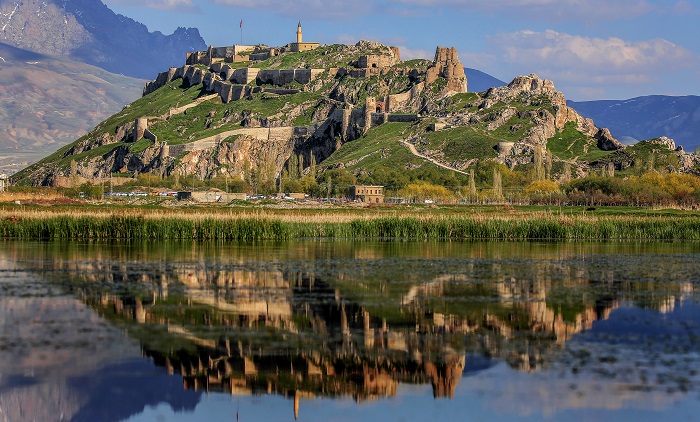 Dağlık alanda yer alan bir kale ve çevresindeki yeşil doğa manzarası, göletteki yansımasıyla birlikte