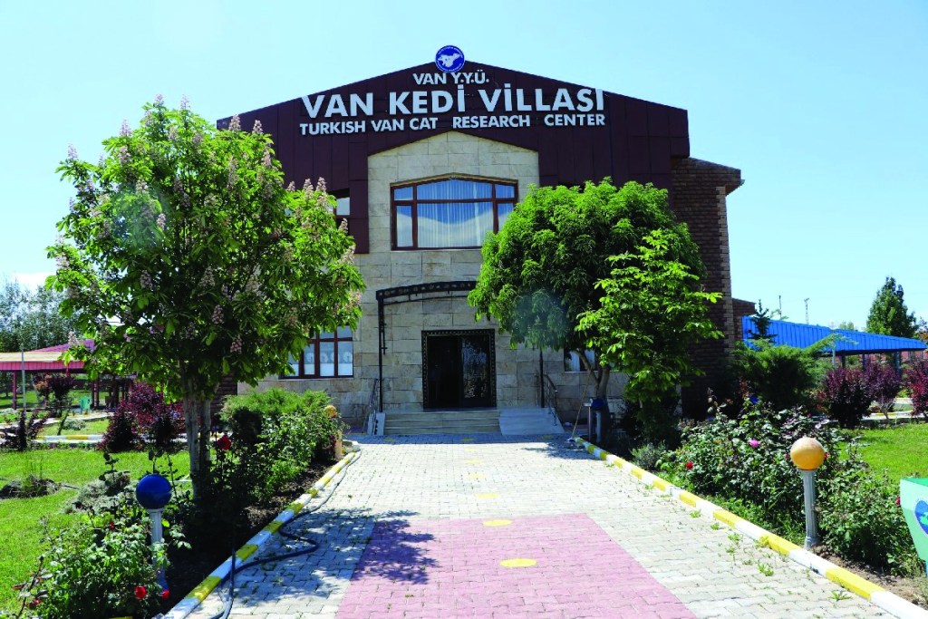 Van Kedi Villası, Türk Van Kedisi Araştırma Merkezi'nin giriş kısmı, çevresinde yeşil ağaçlar ve çiçeklerle dolu bir bahçe.