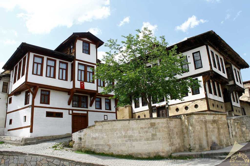 Geleneksel Türk mimarisini yansıtan, beyaz ve kahverengi ahşap detaylarla süslü iki katlı evler ve önünde bir ağaç.