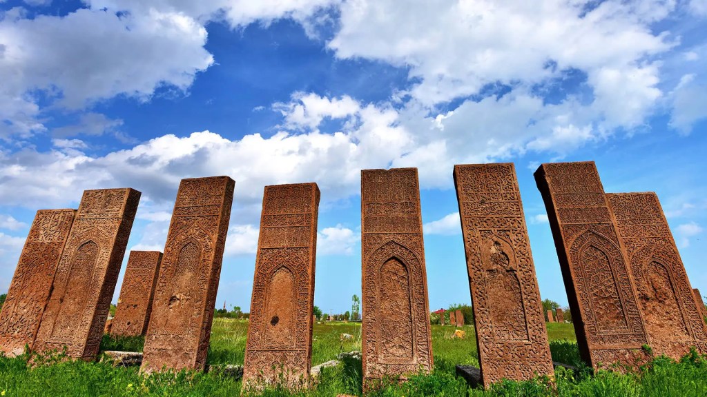 Ahlat Selçuklu Mezarlıkları'nda bulunan, tarihi taş işçiliğiyle süslü mezar taşları, geniş bir yeşil alanda sıralanmış olarak görülüyor. Arka planda mavi gökyüzü ve beyaz bulutlar dikkat çekiyor.