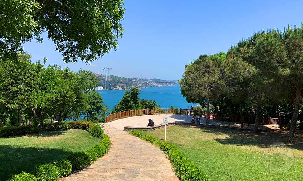 Yeşil ağaçlarla çevrili bir yürüyüş yolu ve Boğaz manzarası, insanların dinlendiği bir alan.