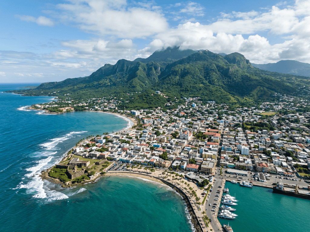 A coastal city with buildings, a harbor, beaches, and green mountains under partly cloudy sky